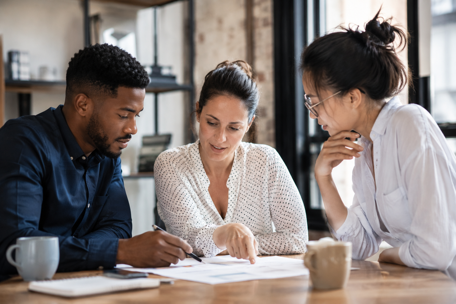 Three colleagues review documents at a wooden table in a bright office setting, collaborating.