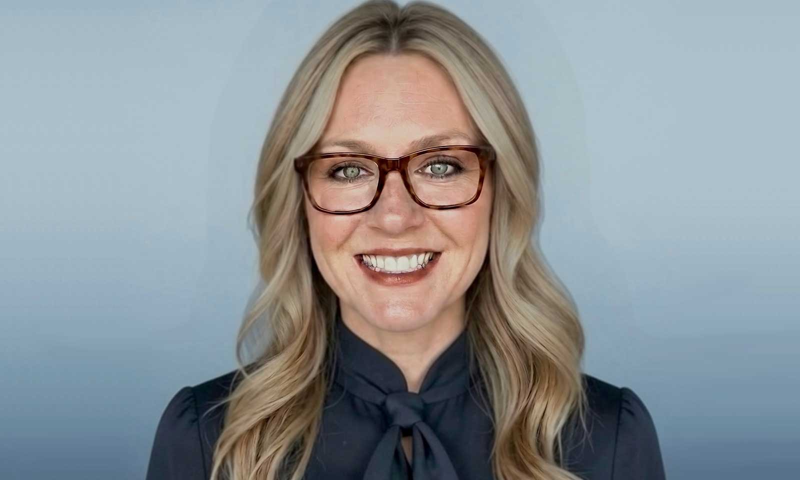 Smiling woman with blonde hair and brown tortoiseshell glasses, wearing a dark blouse with a bow, against a blue backdrop.